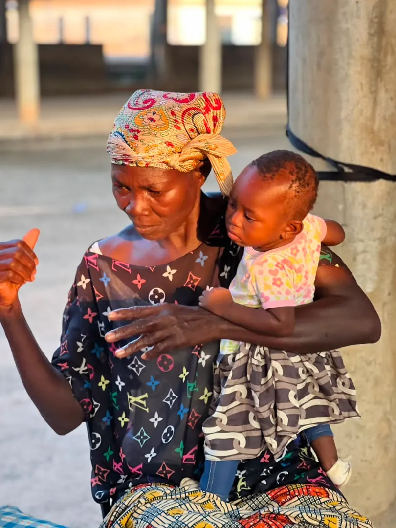 A community member in a colourful headwrap holds a young child outdoors in warm sunlight at the Farmers’ Engagement in Akyawkrom