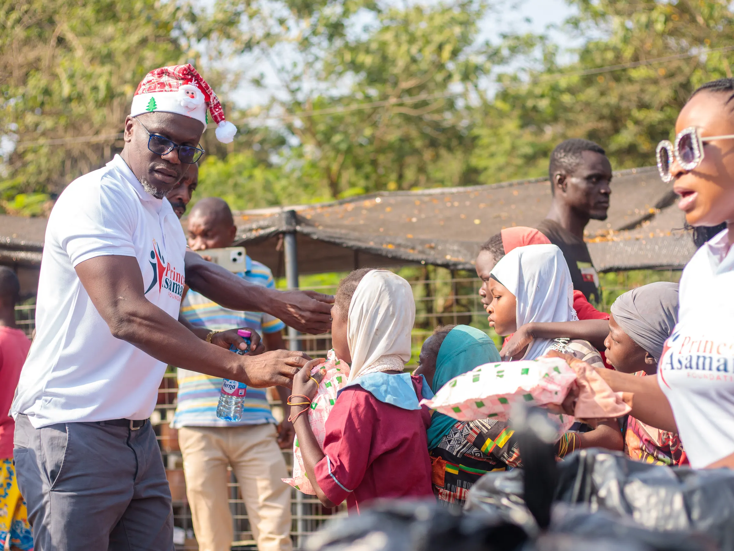 Foundation volunteer in a Santa hat handing a holiday gift to a child over a car boot during the Joy to the Street outreach in Kumasi